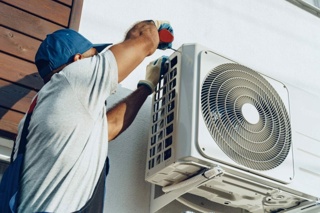 Technician in a blue cap and gloves installing or repairing an outdoor air conditioning unit mounted on a wall in Texas.