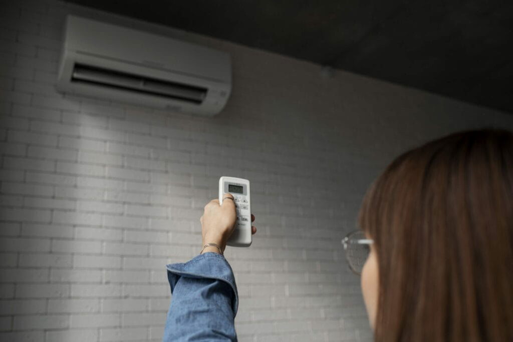 Person pointing a remote control towards an HVAC air conditioner mounted on a wall inside a room with white brick walls in Texas.