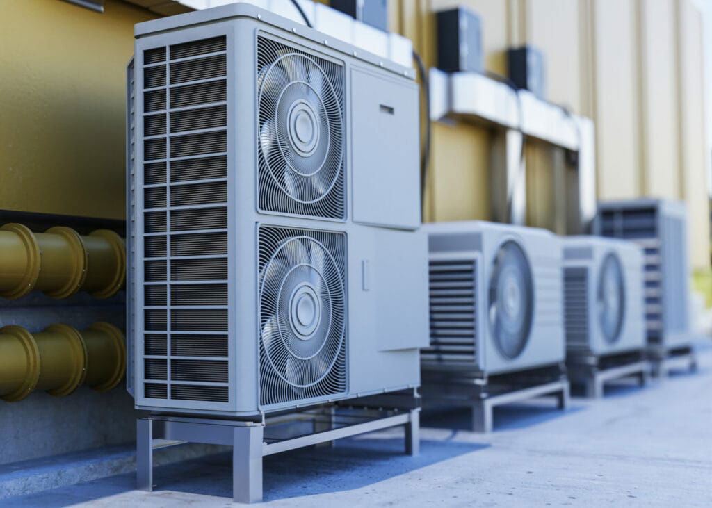 Outdoor HVAC units with large fans are installed in a row next to a building in Texas. The metallic units are elevated on metal stands with yellow pipes visible on the left side.
