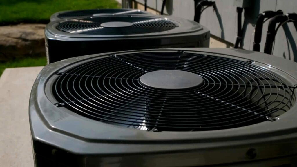 Close-up of two outdoor air conditioning units with black fan grills, positioned side by side against a building wall. Green grass is visible in the background. Patriot Appliance Repair & HVAC services in Austin ensure your HVAC systems run efficiently throughout Texas.