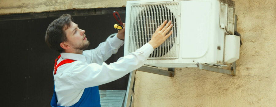A technician in a blue overall adjusts an outdoor HVAC unit mounted on a wall, using tools, with a screwdriver and drill visible near the unit.